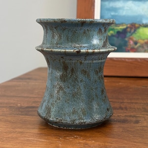 May include: A ceramic vase with a flared rim and a central horizontal ridge. The piece features a mottled glaze in shades of dusty blue and dark brown, creating a textured appearance across the entire surface of the cylindrical vessel.