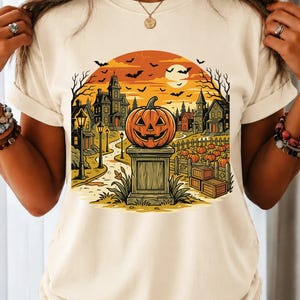 May include: A cream-colored t-shirt featuring a graphic print of a carved jack-o'-lantern sitting on a stone pedestal. The scene includes a spooky village with houses, bare trees, a full moon, and flying bats against an orange sunset sky.