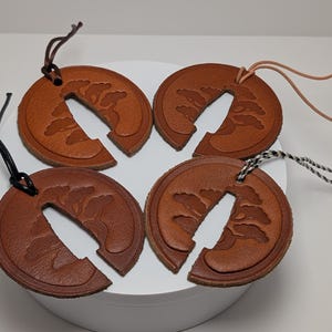Peut inclure: Quatre étiquettes circulaires en cuir marron avec un motif d'arbre bonsaï en relief et une fente verticale centrale. Chaque étiquette comprend un cordon fin pour la suspension, avec des variations de couleur dont noir, marron foncé, beige et torsadé.