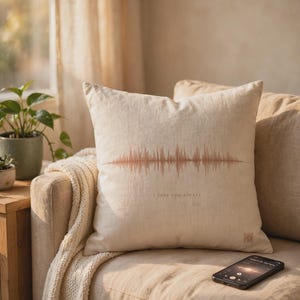 Peut inclure: Un coussin carré beige orné d'un graphique d'onde sonore de couleur cuivre imprimé au centre. Sous la forme d'onde, le texte "i love you always" est inscrit dans une petite police simple. Le coussin présente un aspect texturé, semblable à du lin.