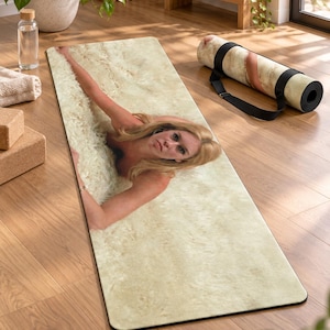 May include: A rectangular yoga mat featuring a photographic print of a person with blonde hair lying on a textured, cream-coloured surface. The mat has rounded corners and a smooth surface, with a matching rolled version nearby featuring a black carry strap.