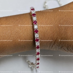 May include: A silver-toned tennis bracelet featuring an alternating pattern of round, vibrant red stones and clear, sparkling white stones set in a continuous line. The bracelet is secured around a wrist, showcasing its flexible, linked metal design.
