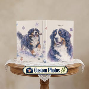 Puede incluir: Un libro de tapa dura con una ilustración de un perro de montaña de Berna en la portada. El frente muestra al perro corriendo en la nieve con un palo, mientras que el reverso presenta un retrato con el nombre Baxter y huellas moradas.