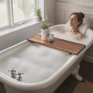 Peut inclure: Un plateau de baignoire rectangulaire en bois posé sur les bords d'une baignoire blanche à pieds remplie de mousse. Le plateau supporte une petite plante en pot blanc et un bougeoir en verre gris. Le bois présente un grain naturel.