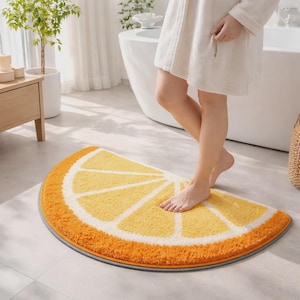 Può includere: Un tappetino da bagno semicircolare con un motivo a spicchi di agrumi giallo e arancione vivace e un bordo bianco. Il tappeto presenta una superficie morbida e testurizzata con una finitura grigia, simile a una fetta di limone o arancia.