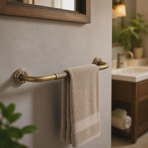 Può includere: Un portasciugamani da parete con finitura in ottone antico spazzolato e piastre di montaggio circolari decorative con un motivo a volute in rilievo. Un asciugamano in cotone beige chiaro piegato è appeso alla barra metallica.