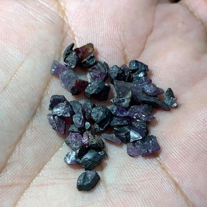 Könnte beinhalten: Eine Ansammlung kleiner, roher und unpolierter Edelsteinfragmente in einer Mischung aus tiefem Anthrazitgrau und durchscheinenden violetten Farbtönen. Diese unregelmäßigen, gezackten Mineralstücke weisen eine natürliche, raue Textur auf.