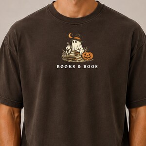 May include: A charcoal grey short-sleeve t-shirt featuring a graphic of a ghost wearing a witch hat, holding a steaming mug, sitting by a stack of books and a jack-o'-lantern. The text "BOOKS & BOOS" is printed in white below the illustration.