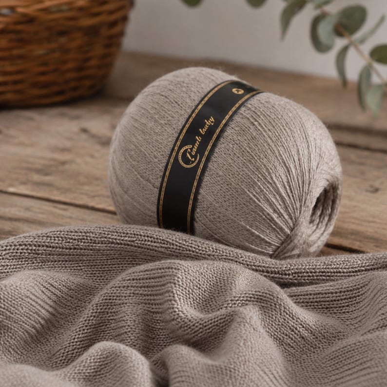 May include: A ball of light brown yarn wrapped in a black paper band with gold text that reads "Lamb baby". The yarn is textured and soft, with a portion of the same light brown knitted fabric draped in front of the ball, showing a consistent stitch.