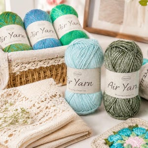 May include: Several skeins of Air Yarn in shades of green, blue, and turquoise are arranged in and around a woven basket. The yarn labels feature the text Air Yarn and Wearing a better life. A crocheted square and a lace-trimmed fabric are also visible.