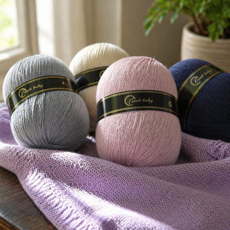May include: Four round balls of yarn in shades of gray, cream, light pink, and navy blue. Each ball features a black paper band with gold trim and the text Lamb baby printed in gold. The yarn balls rest on a textured light purple knitted fabric.