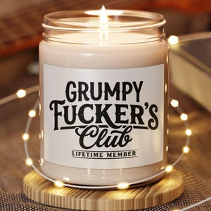 May include: A lit candle in a clear glass jar with a white label featuring the black text "GRUMPY FUCKER'S Club" and "LIFETIME MEMBER" in a stylised font. The candle sits on a round wooden coaster, surrounded by thin, glowing string lights.