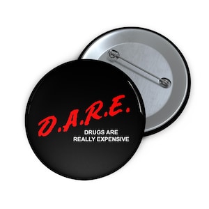 May include: A round black pin-back button featuring the red text D.A.R.E. and the white text DRUGS ARE REALLY EXPENSIVE. The back of the button is silver-coloured metal with a pin mechanism visible on the second button positioned behind the first.