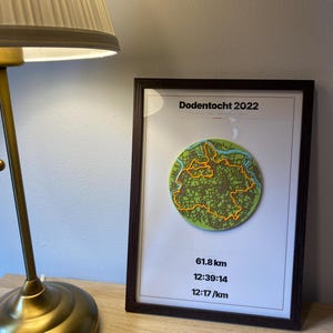 Puede incluir: Una pieza de arte mural enmarcada titulada Dodentocht 2022 que presenta un mapa circular verde con una línea de ruta naranja. Debajo del mapa, el texto muestra 61,8 km, 12:39:14 y 12:17 /km. La impresión está en un marco marrón oscuro con un borde blanco.