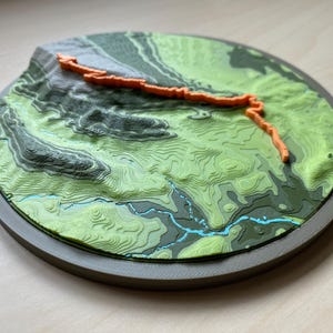 Może przedstawiać: Okrągła mapa topograficzna 3D z warstwowymi odcieniami zieleni przedstawiającymi wysokość, z jaskrawopomarańczową wypukłą ścieżką szlaku i cienkimi niebieskimi liniami odwzorowującymi strumienie, zamknięta w solidnej ciemnoszarej okrągłej ramie.