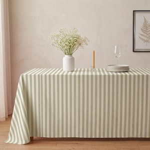 May include: A rectangular tablecloth featuring a vertical striped pattern in alternating shades of sage green and white. The fabric drapes over the edges of a table, providing a clean, uniform look for a dining surface with a classic textile design.