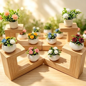 Könnte beinhalten: Eine Sammlung von künstlichen Miniatur-Blumenarrangements in kleinen, gerippten weißen Keramiktöpfen, präsentiert auf abgestuften hellen Holzständern. Die Blumen haben verschiedene Farben wie Rosa, Gelb, Blau, Weiß und Rot mit grünen Blättern.