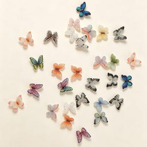 May include: A collection of small, translucent butterfly-shaped charms in various colors including blue, orange, pink, white, grey, and green. Each butterfly features delicate wing patterns and a smooth, semi-transparent finish, arranged loosely.