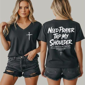 May include: A charcoal gray V-neck t-shirt featuring a white cross graphic on the front left chest. The back displays the white text "NEED PRAYER TAP MY SHOULDER" followed by "LET'S TAKE IT TO GOD TOGETHER" and a small cross symbol.