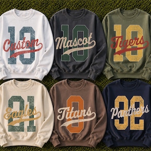 May include: Six crewneck sweatshirts in white, charcoal, olive, cream, brown, and navy. Each features a large distressed number graphic with a script word overlay: Custom, Mascot, Tigers, Eagles, Titans, and Panthers in various colors.