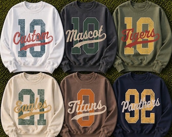 May include: Six crewneck sweatshirts in white, charcoal, olive, cream, brown, and navy. Each features a large distressed number graphic with a script word overlay: Custom, Mascot, Tigers, Eagles, Titans, and Panthers in various colors.