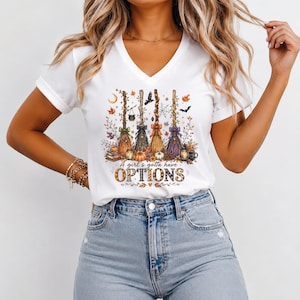 May include: A white V-neck t-shirt featuring a graphic print of four colorful witch brooms, autumn leaves, pumpkins, and a small cauldron. The text "A girl's gotta have OPTIONS" is printed below the brooms in a decorative, patterned font.