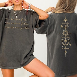 May include: A charcoal gray short-sleeve t-shirt featuring gold text on the front reading YOU DO NOT fear, YOU DO NOT falter, YOU DO NOT yield. The back displays a vertical gold graphic with a lotus, sun, moon, and butterflies with text.