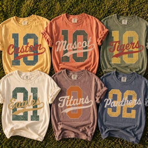 May include: Six short-sleeve crew neck t-shirts in yellow, orange, olive green, cream, mauve, and blue. Each shirt features a large distressed-style number graphic with a script word overlaid, including Custom, Mascot, Tigers, Eagles, Titans, and Panthers.