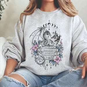 May include: A light gray crewneck sweatshirt featuring a detailed black ink illustration of a dragon perched on a stack of books. The design includes purple flowers, a mountain landscape, moon phases, and the text: Only you can decide what breaks you.