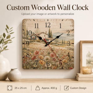 May include: A square wooden wall clock measuring 25 x 25 cm with a weight of approximately 400 g. The clock face features a printed landscape illustration of a house in a field of flowers, black numerals, and black hands with a red second hand.