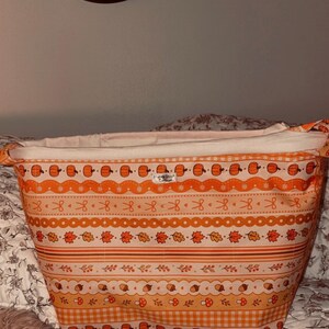 May include: A fabric storage basket featuring a horizontal pattern of orange pumpkins, autumn leaves, acorns, and mushrooms on a light background. The basket has a white interior lining and a small rectangular label reading Honey Stitch Cottage.
