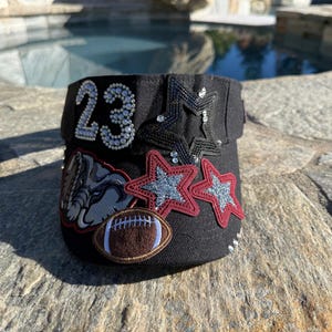 May include: A black visor featuring the number 23 in silver rhinestones, a black sequined star, two red and silver glitter stars, a brown football patch, and a grey bulldog mascot patch. The visor is decorated with various embellishments.