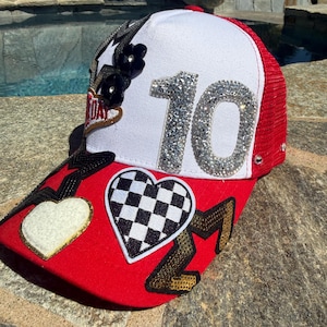 May include: A red and white trucker hat featuring a large rhinestone number 10 on the front panel. The red brim is decorated with a white heart patch, a black and white checkered heart patch, and a gold sequined star patch. Black flower accents.