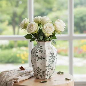 May include: A bouquet of cream-coloured roses with green leaves arranged in a white ceramic vase. The vase features a hexagonal shape and is decorated with a delicate, repeating pattern of green vines, small leaves, and tiny brown floral accents.