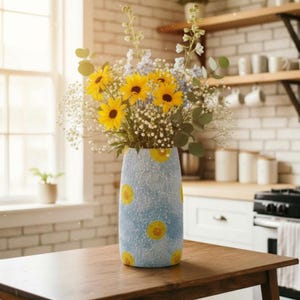 May include: A cylindrical vase with a textured light blue surface featuring yellow sunflower patterns, holding a bouquet of bright yellow sunflowers, small white baby's breath flowers, and tall green stems with light blue blossoms.