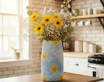 May include: A cylindrical vase with a textured light blue surface featuring yellow sunflower patterns, holding a bouquet of bright yellow sunflowers, small white baby's breath flowers, and tall green stems with light blue blossoms.