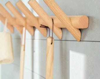 May include: A wall-mounted wooden rack featuring five angled pegs extending from a horizontal base. The light-toned wood has a natural grain pattern. Several kitchen utensils with long wooden handles are hanging from the pegs by dark cords.