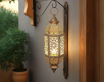 May include: A hanging metal lantern with a distressed silver finish and intricate geometric cutouts. The lantern is suspended from a black wrought iron wall bracket and emits a warm, glowing light from within its hexagonal frame.