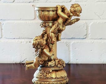 May include: A gold-coloured candle holder featuring a central pillar adorned with two cherub figures. The base and the top cup are decorated with sculpted rose motifs and fluted detailing. The entire structure has a metallic finish.