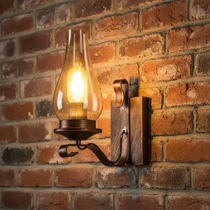 Puede incluir: Un aplique de pared con base de metal en tono bronce y un brazo curvo que sostiene una pantalla de cristal transparente en forma de lágrima. La lámpara contiene una bombilla encendida que emite una luz cálida y brillante.