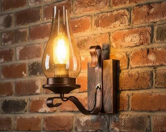 May include: A wall-mounted light fitting featuring a bronze-toned metal base and a curved arm supporting a clear, teardrop-shaped glass shade. The fitting contains a glowing light bulb that emits a warm, bright illumination from within.