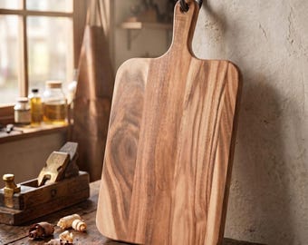 May include: A rectangular wooden chopping board with a rounded handle and a hole for hanging. The board features a natural light and dark brown wood grain pattern with smooth, rounded edges, displayed in an upright position.