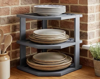 May include: A three-tiered grey corner shelf unit constructed with a triangular frame. The top shelf holds a stack of white plates, the middle shelf holds a stack of light-coloured plates, and the bottom shelf holds a stack of tan plates.