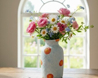 May include: A light blue textured vase with orange and yellow circular spots containing a bouquet of pink roses, white daisies, and small blue flowers. The vase has a cylindrical shape with a slightly tapered neck and a rough, matte finish.