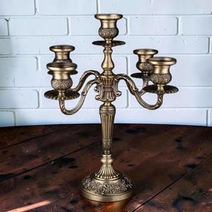 May include: A five-arm candelabra crafted from antique brass-toned metal, featuring intricate floral embossed detailing on the base and candle cups, a central stem with vertical fluting, and curved arms supporting each individual candle holder.