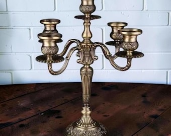 May include: A five-arm candelabra crafted from antique brass-toned metal, featuring intricate floral embossed detailing on the base and candle cups, a central stem with vertical fluting, and curved arms supporting each individual candle holder.