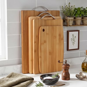 May include: A set of four rectangular bamboo cutting boards of varying sizes arranged vertically. The three largest boards feature silver-toned metal handles at the top, while the smallest board has a circular hole for hanging or gripping.