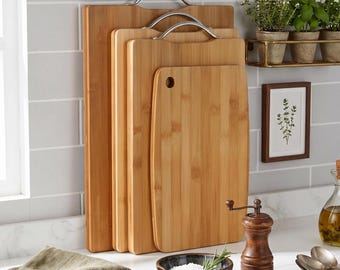 May include: A set of four rectangular bamboo chopping boards of varying sizes arranged vertically. The three largest boards feature silver-toned metal handles at the top, while the smallest board has a circular hole for hanging or gripping.