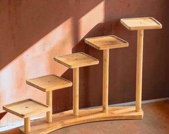 May include: A tiered bamboo plant stand featuring four square platforms of varying heights arranged in a stepped, ascending formation. The light brown wooden structure is supported by a curved base and vertical cylindrical posts for each level.