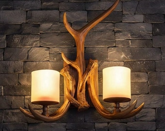 May include: A wall-mounted light fitting featuring a brown, textured antler-shaped frame that supports two cylindrical, frosted glass shades. The design includes a central vertical antler element with two side branches holding the light bulbs.